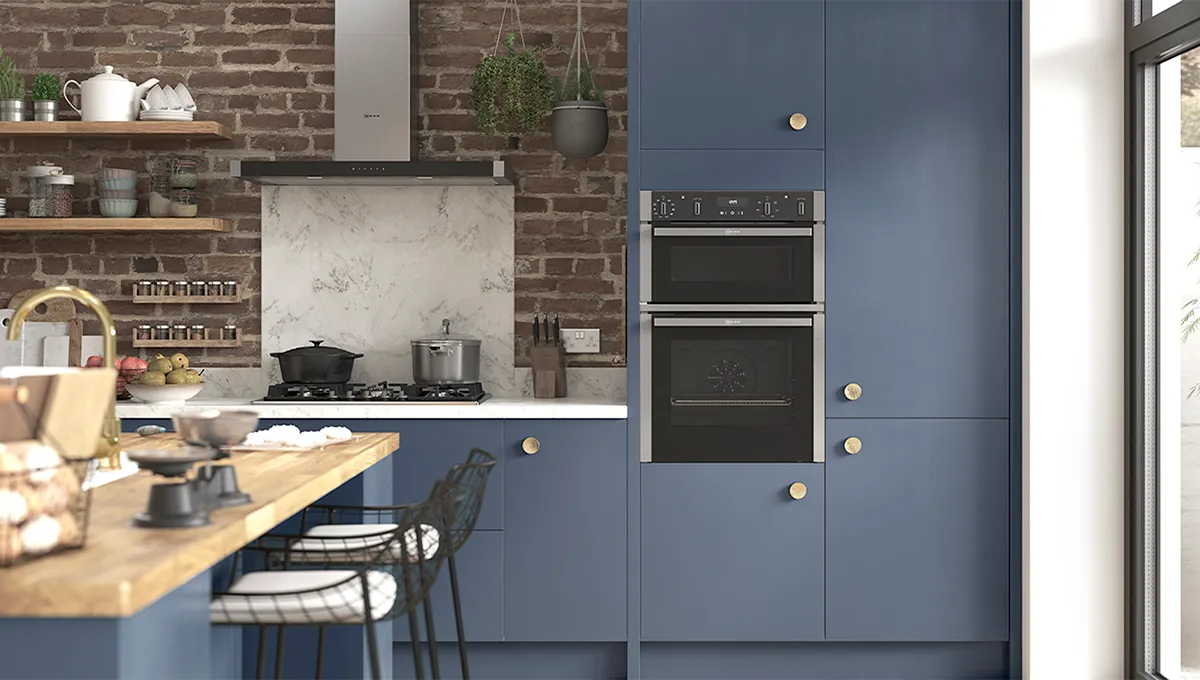A modern kitchen with industrial-chic style, featuring blue cabinets, a built-in stack of black and steel NEFF ovens, and a stainless steel range hood against an exposed brick wall.