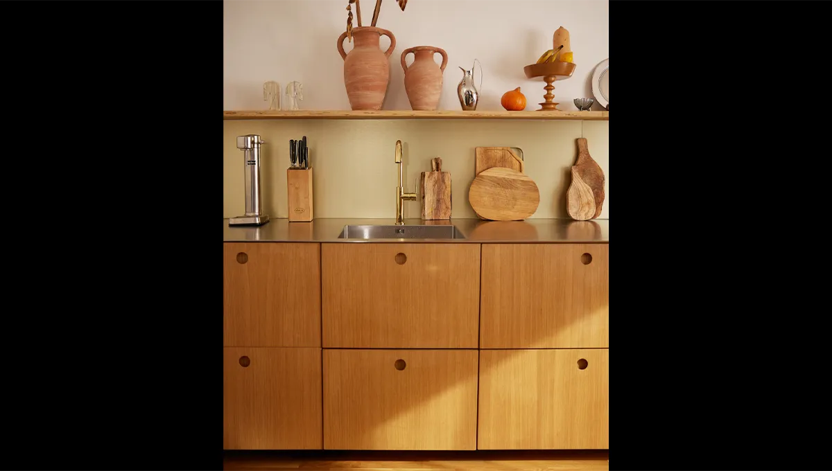 A front-on view of minimalist wooden kitchen cabinets with a stainless steel sink, gold faucet, and a decorative open shelf above.
