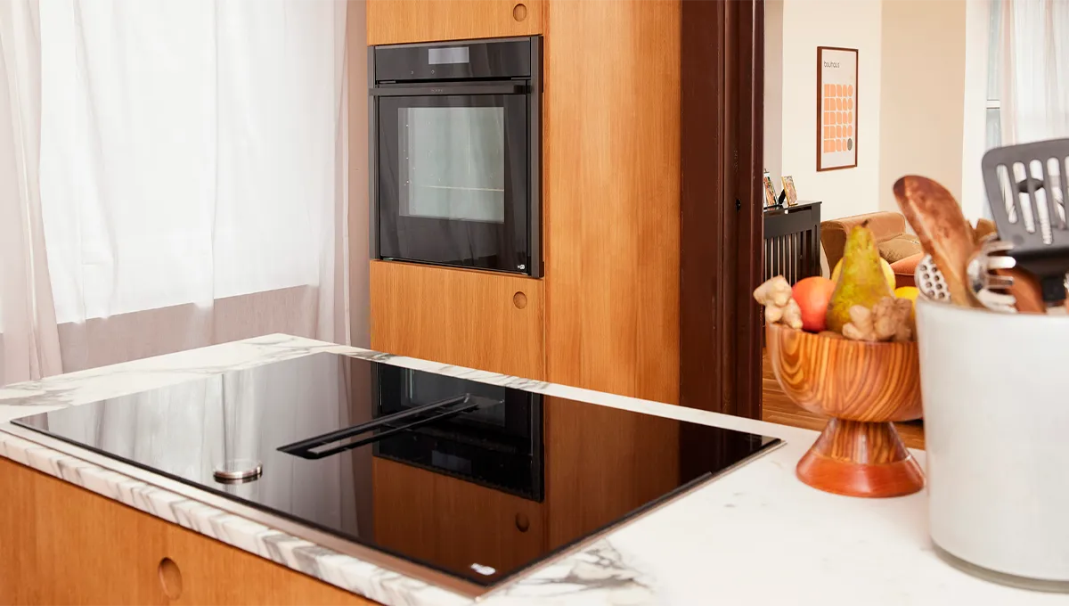 A view across a marble kitchen island with a NEFF vented hob, looking towards a built-in NEFF oven seamlessly integrated into tall wood cabinets.