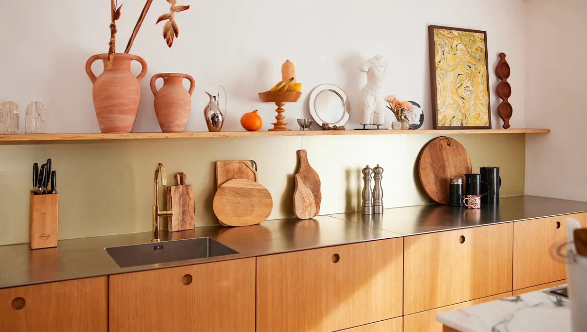A stylish kitchen with light wood cabinets, a stainless steel countertop and sink, and a long open shelf decorated with art and terracotta vases.