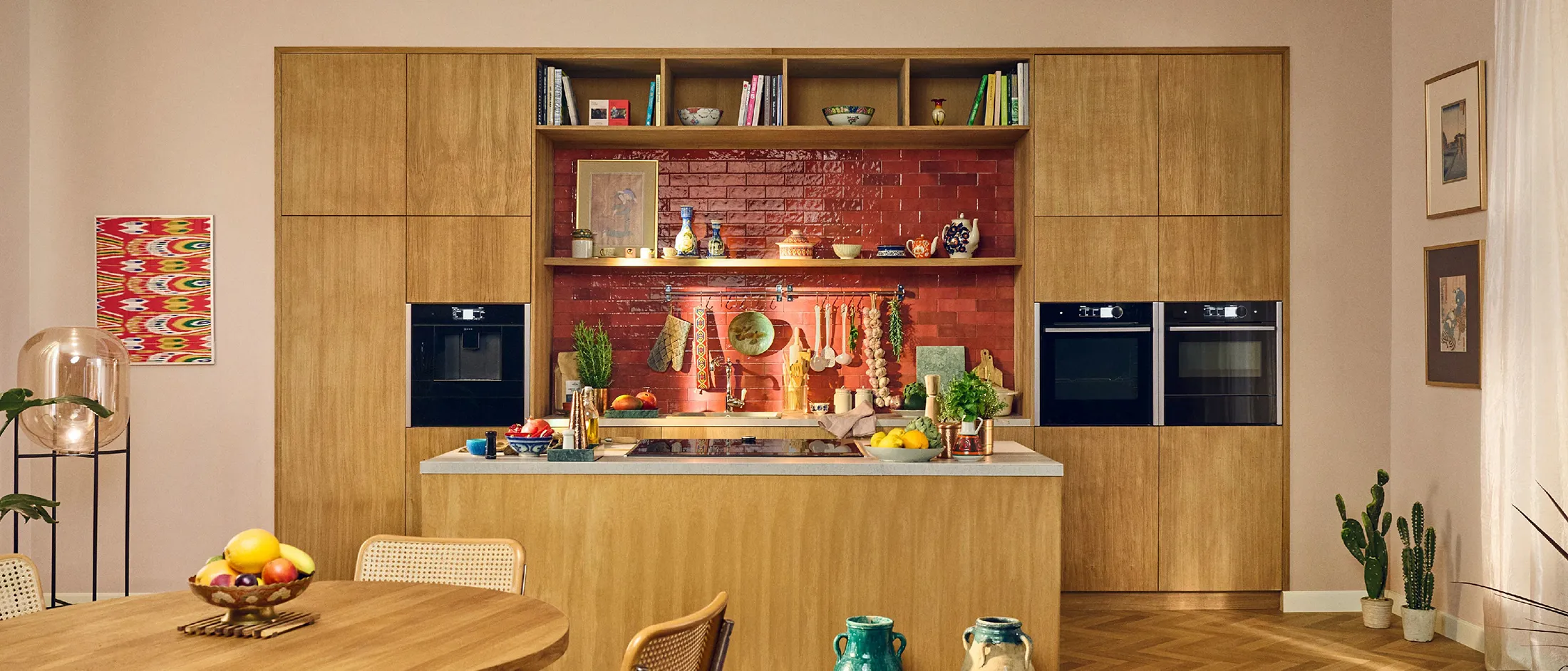 The light oak kitchen with wooden cabinets, red tile backsplash, kitchen island with fruits, plants, and hanging utensils, and dining table with wicker chairs.