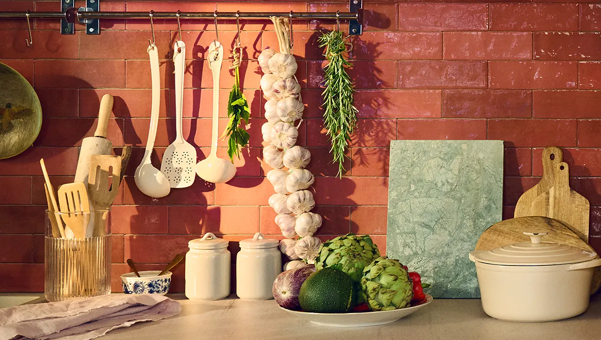 Kitchen counter with dark terracotta backsplash, wooden utensils, hanging garlic and herbs, ceramic jars, fresh vegetables, cutting boards, and a covered pot.