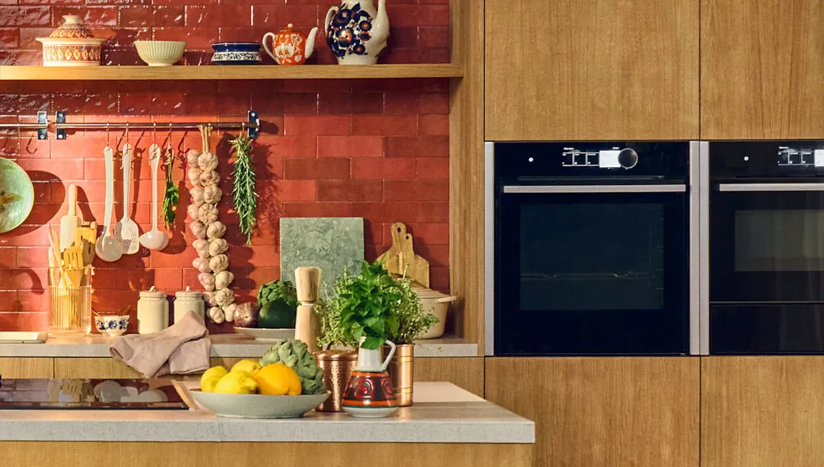Modern kitchen with red tiled backsplash, wooden cabinets, hanging utensils, garlic and herbs, countertop with fruit bowl and potted herbs.
