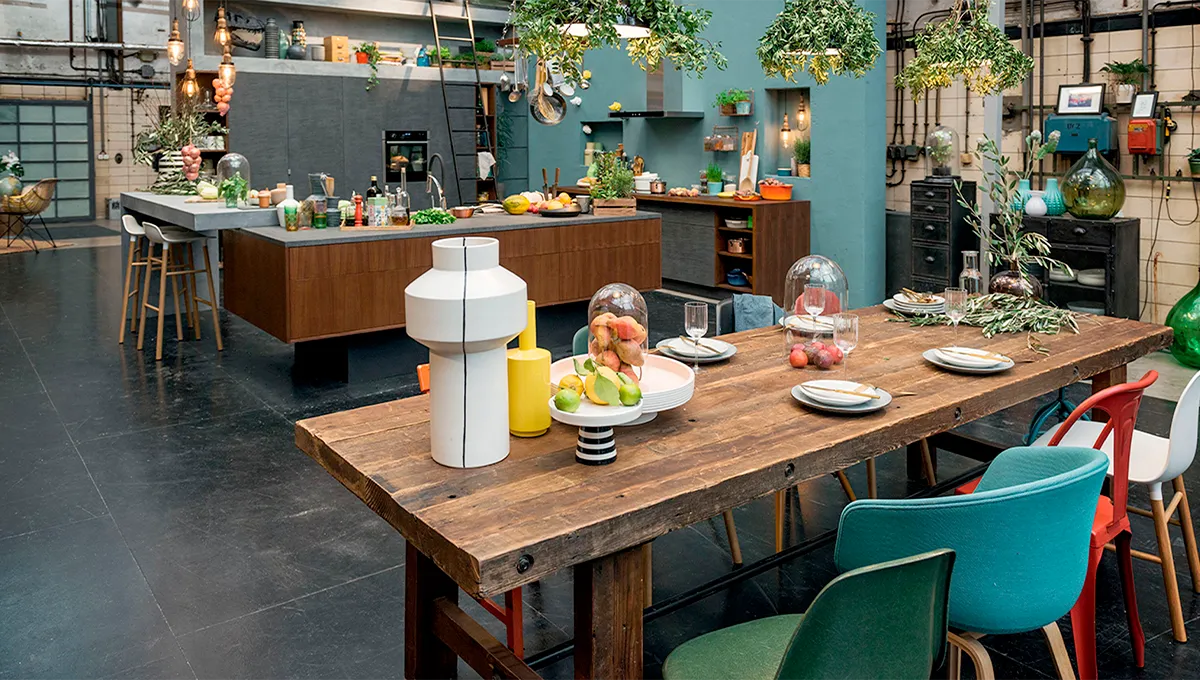 Eine Totalaufnahme eines geräumigen Raums im industriellen Loft-Stil: Im Vordergrund steht ein großer, rustikaler Esstisch aus Holz, der für eine Mahlzeit gedeckt ist und von bunten, nicht zusammenpassenden Stühlen umgeben wird; im Hintergrund befindet sich eine große, moderne Küche.