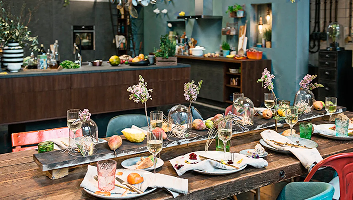 Ein herrlich unordentlicher Esstisch nach einer Mahlzeit – mit Essensresten auf den Tellern, Weingläsern und fantasievollen Blumenarrangements –, der in einem Loft-Ambiente eine warme, festliche Stimmung einfängt.