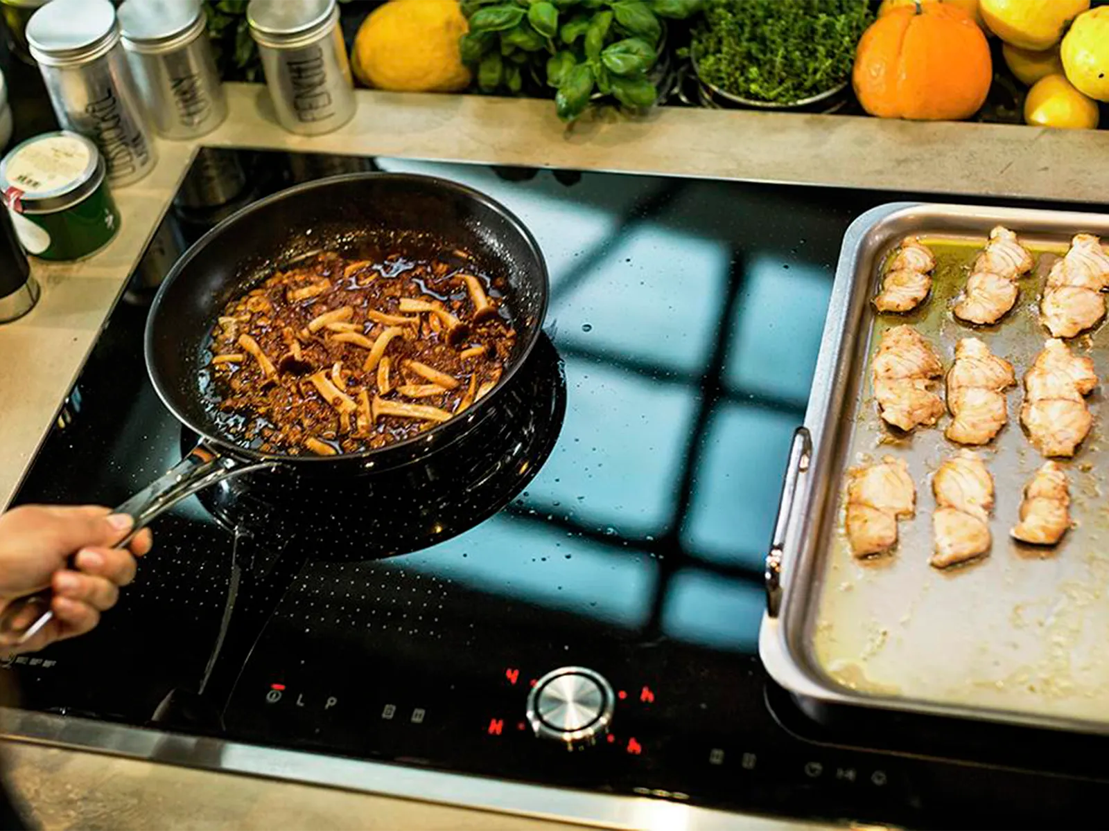 A chef is cooking on a black NEFF induction cooktop, holding a pan with a simmering mushroom sauce, while a tray of seared fish rests beside it, all surrounded by fresh herbs and produce.
