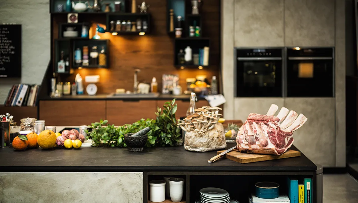 A chef's-eye view of a kitchen island laden with gourmet ingredients, including a large prime rib roast, oyster mushrooms, and fresh herbs, with the NEFF kitchen in the background.