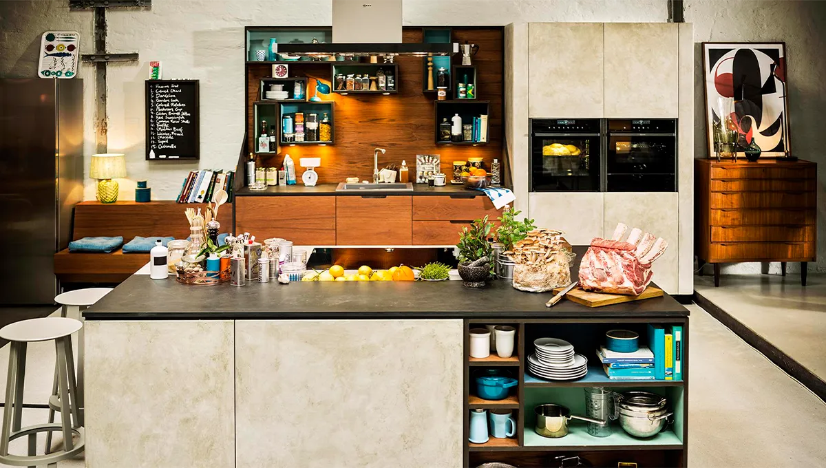A wide shot of an eclectic, high-end kitchen featuring a mix of concrete and wood finishes, a large island with open shelving, and a full suite of NEFF appliances, including two built-in ovens.
