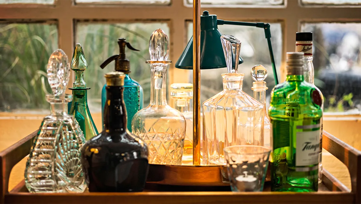 A stylish home bar scene featuring a collection of elegant glass decanters, liquor bottles, and a vintage seltzer bottle arranged on a wooden tray in front of a window.