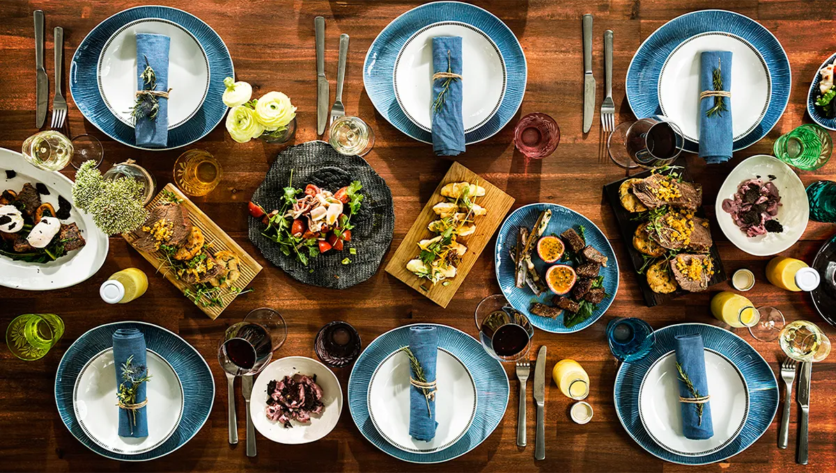 A lavish, top-down view of a beautifully set dining table, abundant with colorful dishes, wine glasses, and stylish blue place settings, ready for a dinner party.