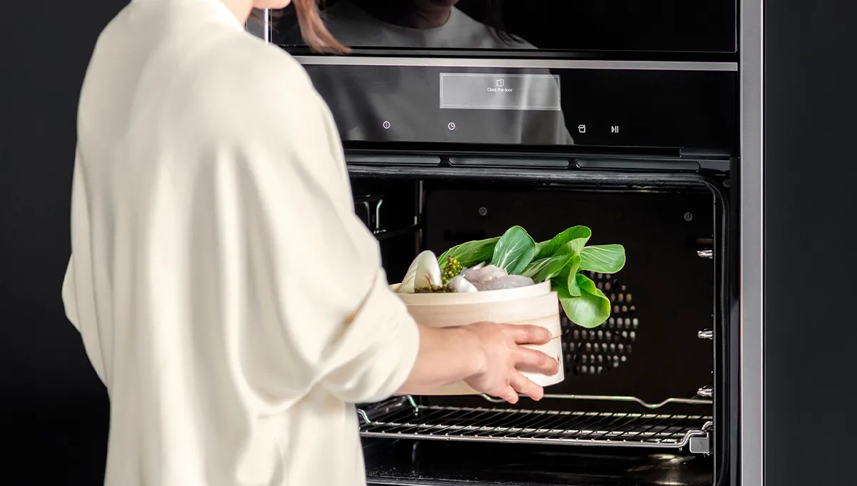 A person placing a bamboo steamer basket filled with fresh pak choi and dumplings into a modern NEFF oven.