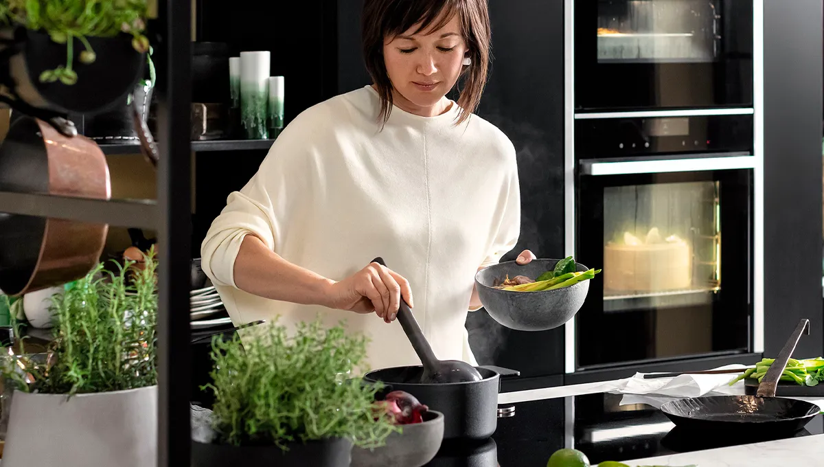 A woman in a modern black kitchen ladles a steaming broth into a bowl, with a NEFF induction cooktop on the island and built-in ovens visible in the background.
