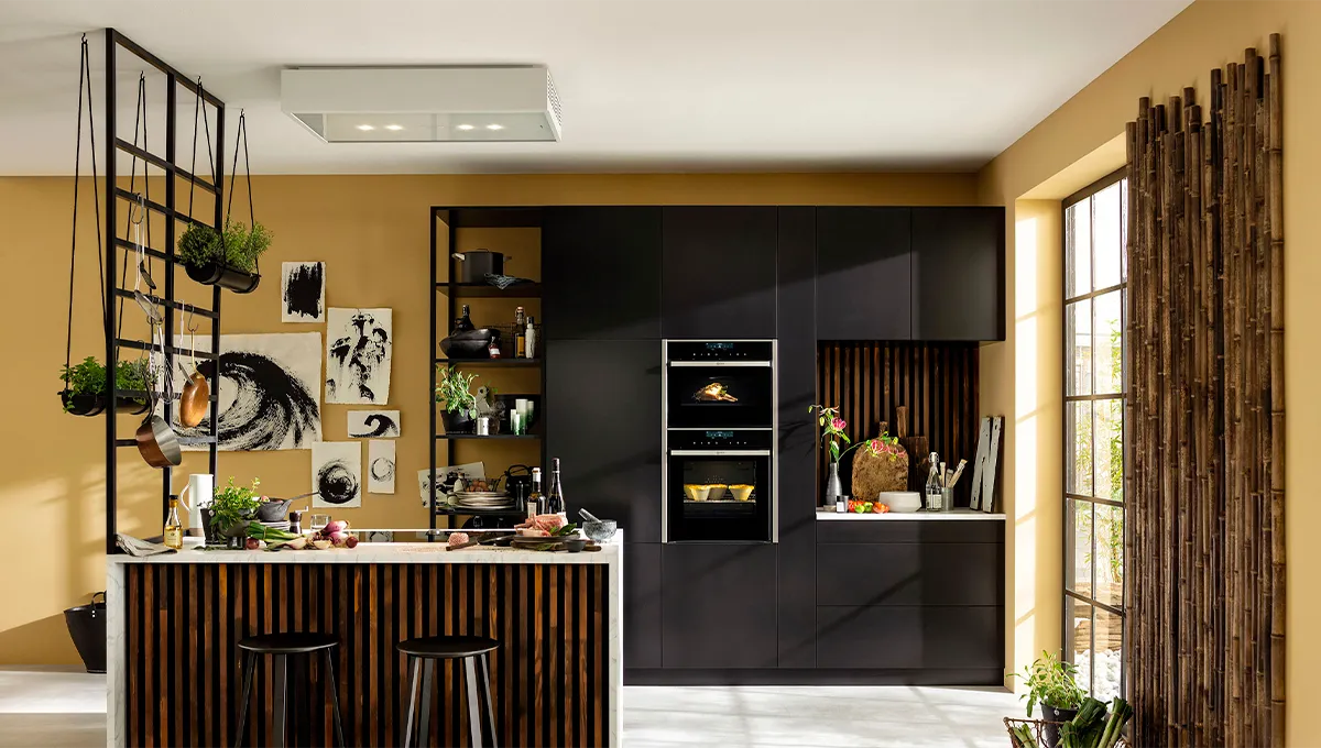 A wide view of a dramatic, modern kitchen with black cabinets, a wood-slatted island, and a vertical stack of black NEFF ovens, accented by a gallery wall and a hanging pot rack.