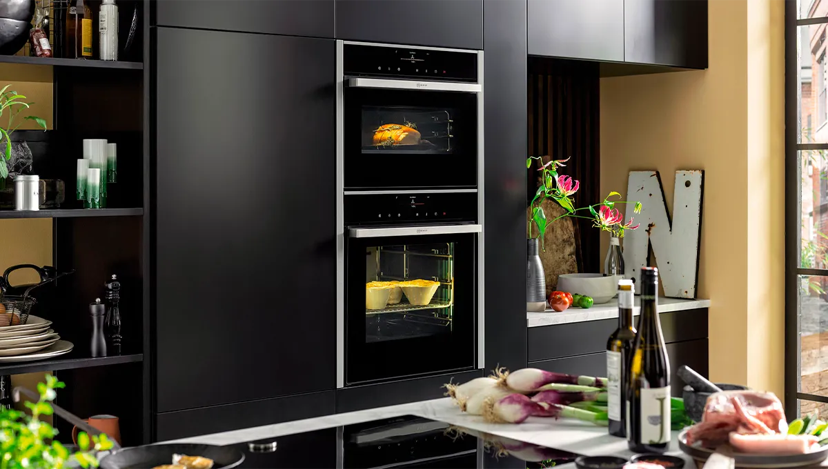 A modern kitchen with matte black cabinetry, showing a vertically stacked pair of NEFF built-in ovens, one with a loaf of bread baking and the other with a casserole dish.