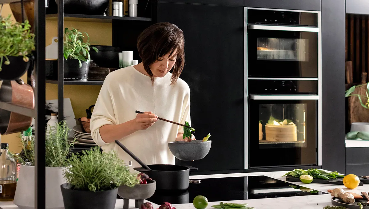 Woman in white sweater cooking with chopsticks in a modern kitchen surrounded by fresh herbs and vegetables.
