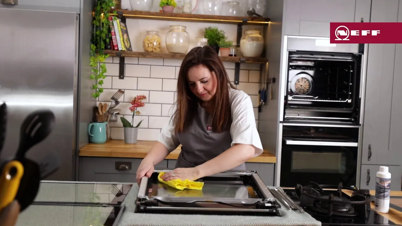 Woman wearing a gray apron and white shirt wiping a glass oven door or stovetop with a yellow cloth on a kitchen counter; modern kitchen cabinets, built-in oven, open shelves with jars and plants, and a stainless refrigerator are visible in the background.