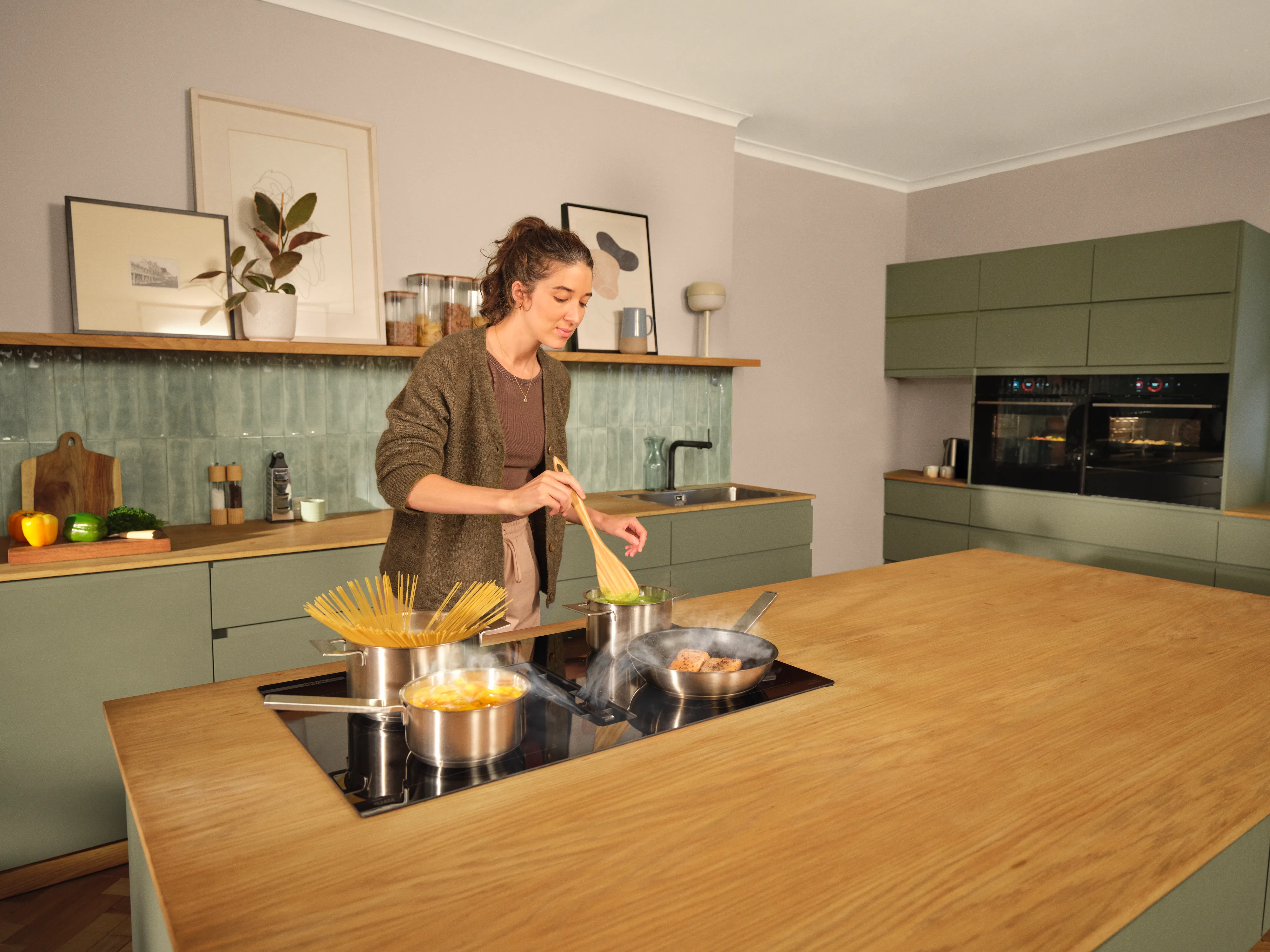 Person cooking at a kitchen island, stirring a pan while a pot of spaghetti boils on an induction cooktop; wooden countertop, sage-green cabinets, built-in ovens and framed artwork on the backsplash.
