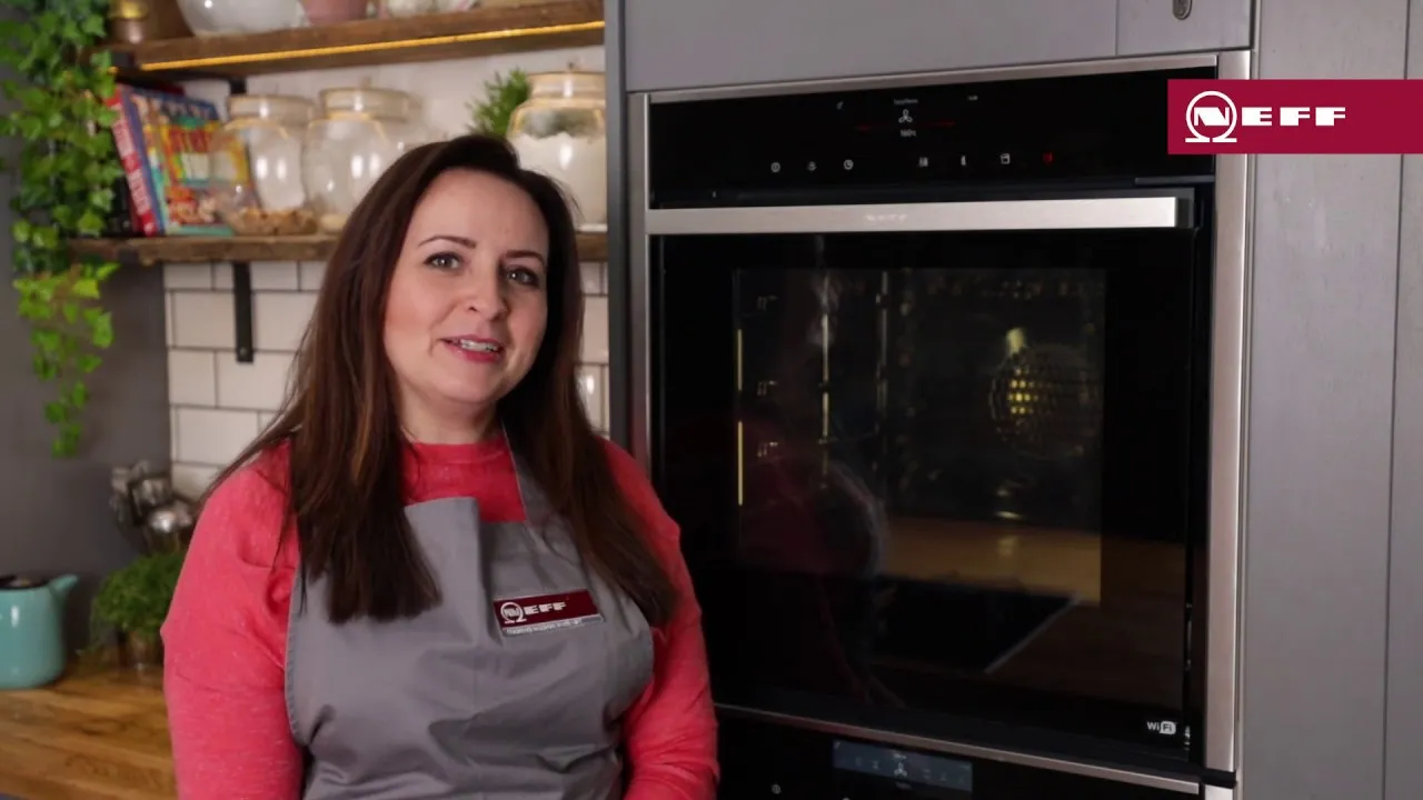 A smiling woman with long brown hair, wearing a gray apron over a red sweater, stands beside a stainless-steel built-in oven in a home kitchen; shelves with cookbooks, jars and a potted plant are visible to the left and a "NEFF" logo appears in the top-right corner.