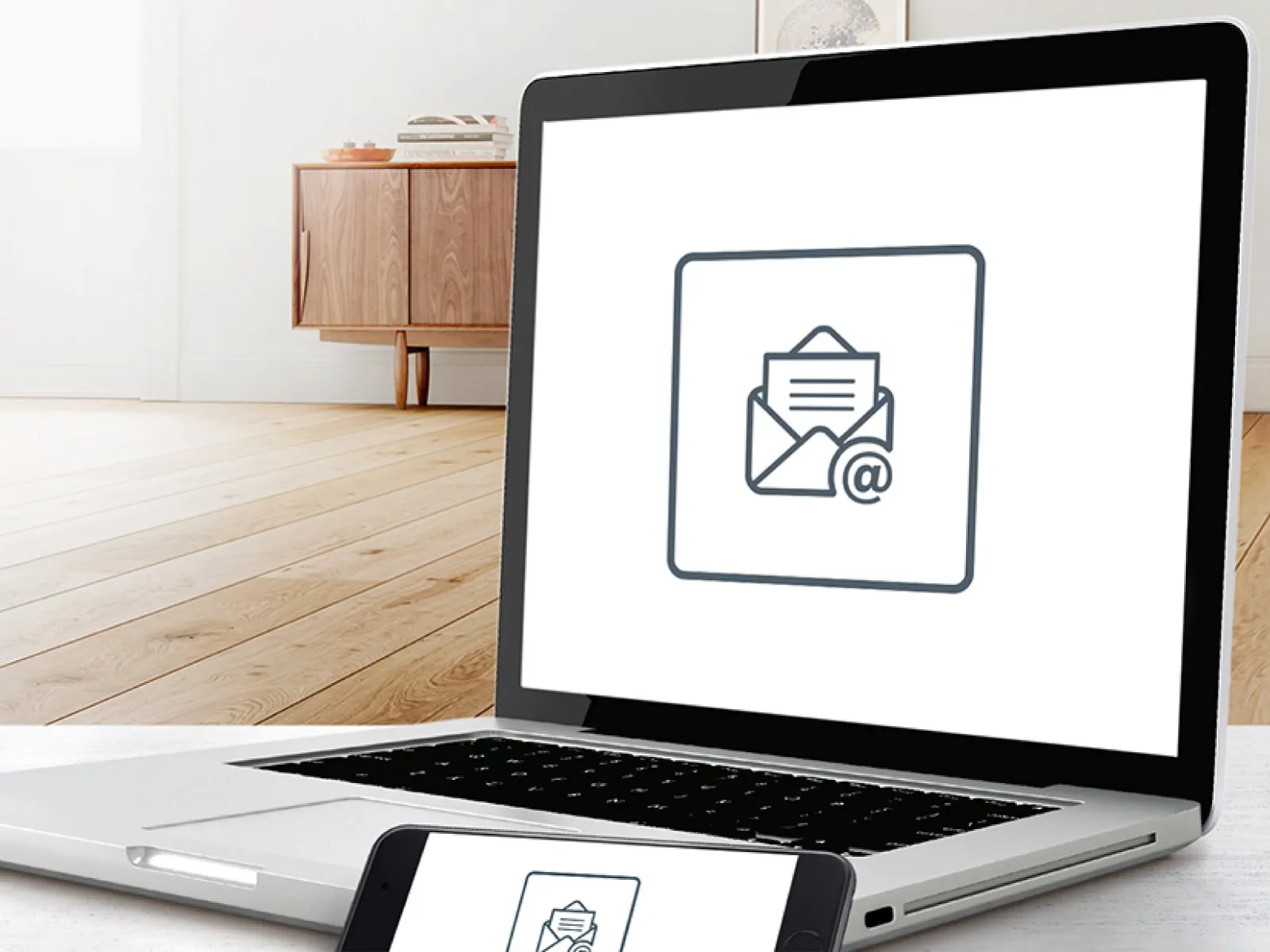 Ein Laptop und ein Handy sind auf einem weißen Tisch in einem Wohnzimmer zu sehen. Auf beiden Geräten erscheint ein Symbol für einen Newsletter.