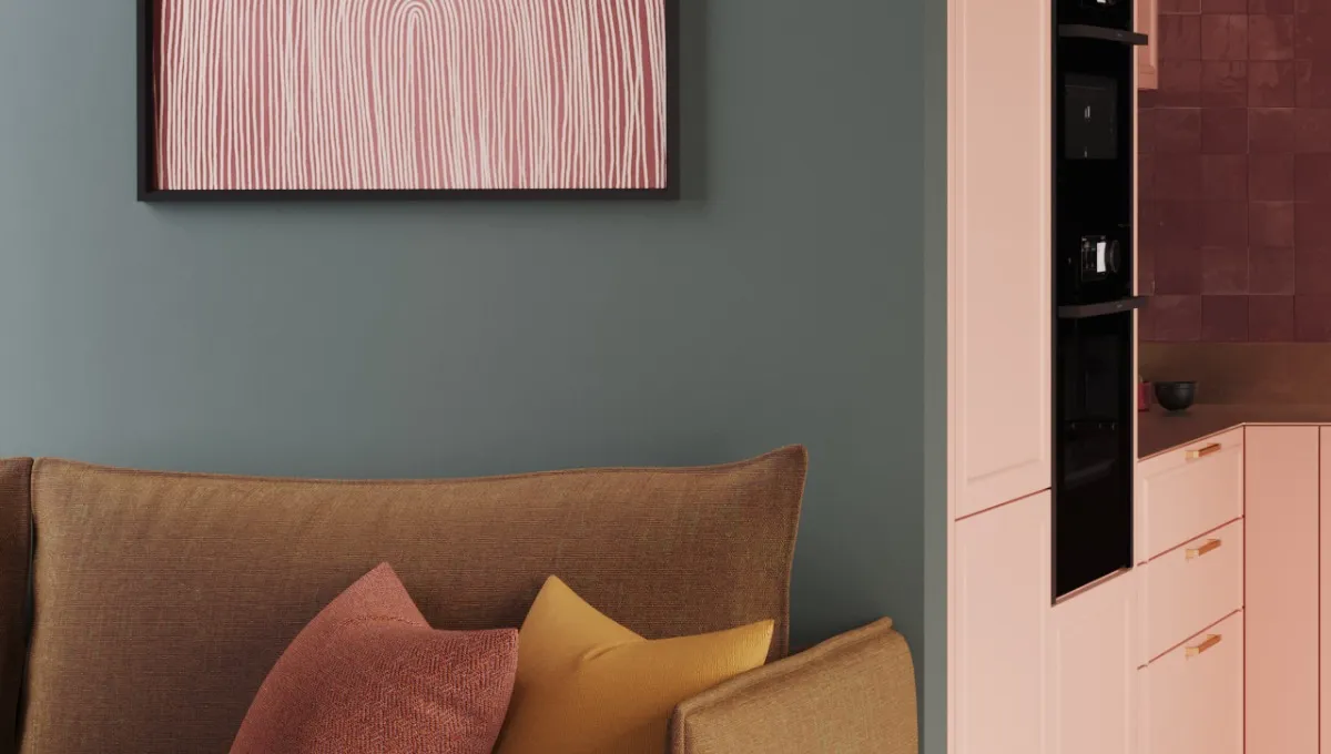 Ein braunes Sofa mit zwei Kissen steht vor einer grünen Wand, an der ein Bild hängt. Im Hintergrund befindet sich eine Küche in einem pfirsichfarbenen Ton mit zwei eingebauten NEFF-Öfen.