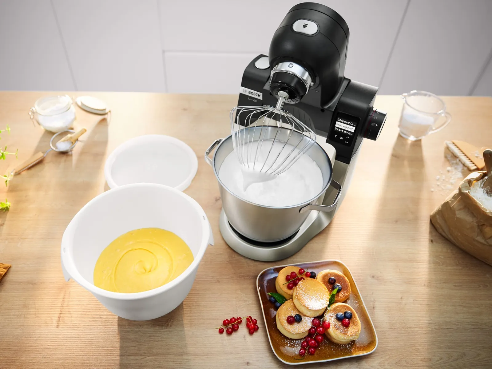 Bosch Series 6 stand mixer whipping batter and cream on a wooden worktop with bowls of batter and mini pancakes topped with berries.