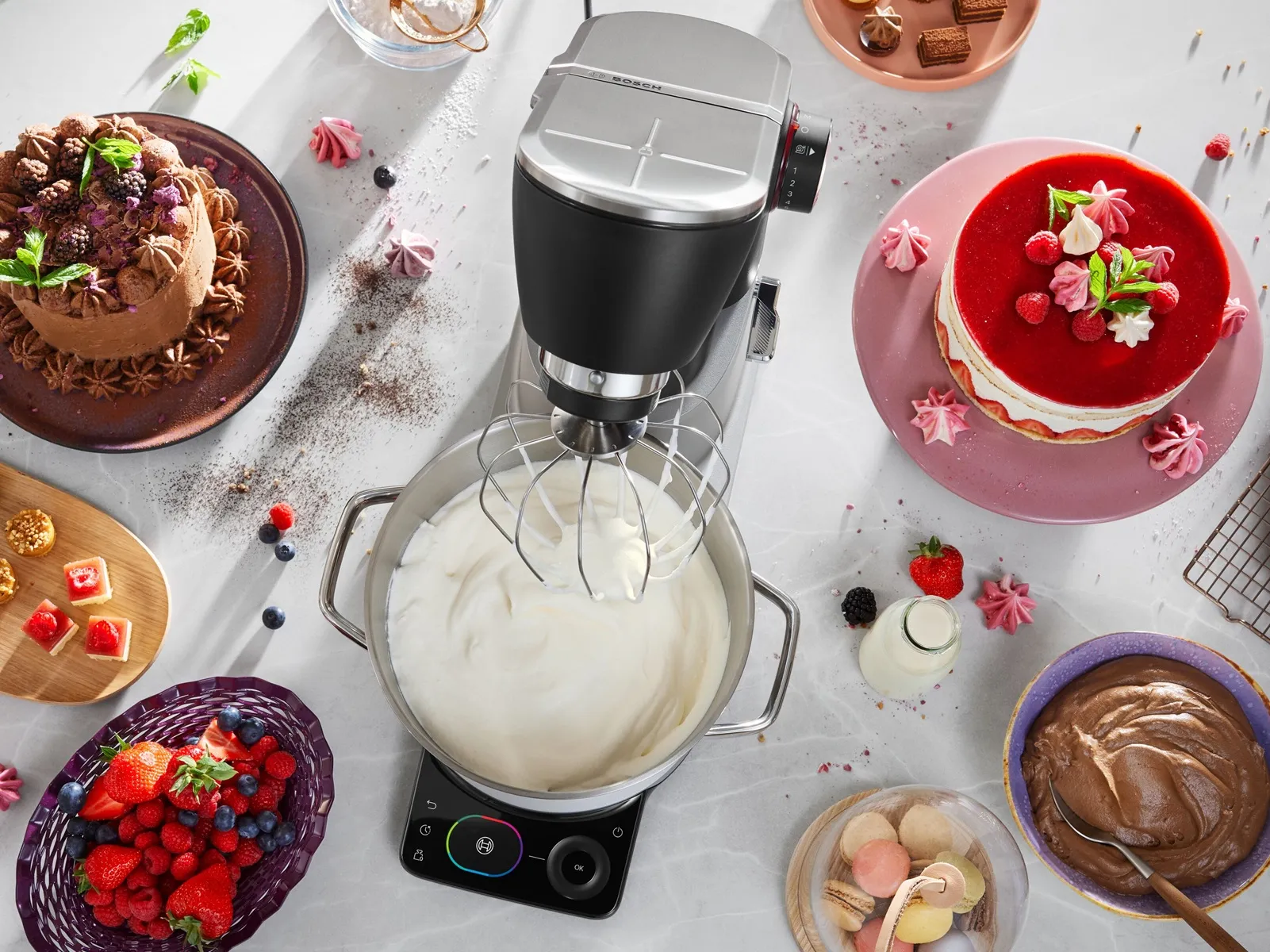 Bosch Series 8 stand mixer whipping cream beside decorated cakes, berries, meringues and chocolate ingredients on a light kitchen worktop.