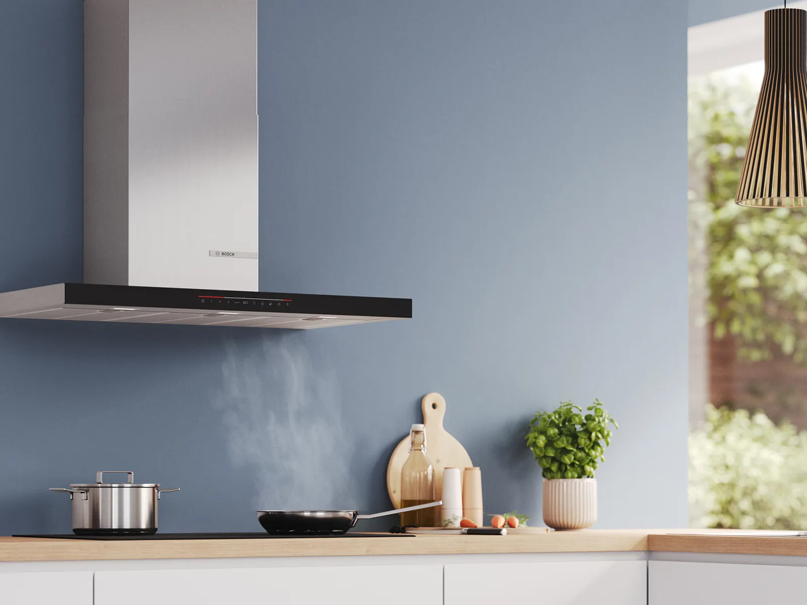 Modern kitchen with a stainless steel range hood, steaming pan, pot, cutting board, herbs, and natural light from a window.