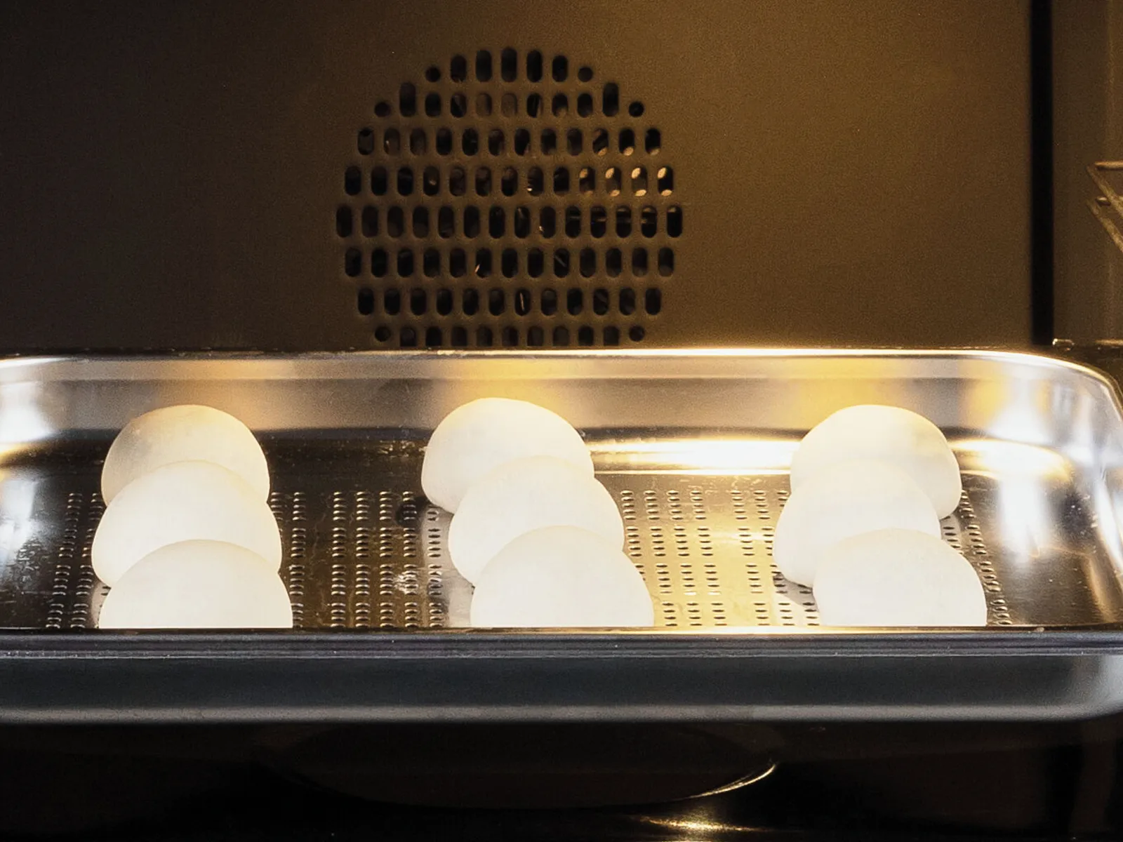Ein gelochtes Backblech mit Reihen von Teiglingen zum Dämpfen in einem modernen, von oben beleuchteten Backofen.