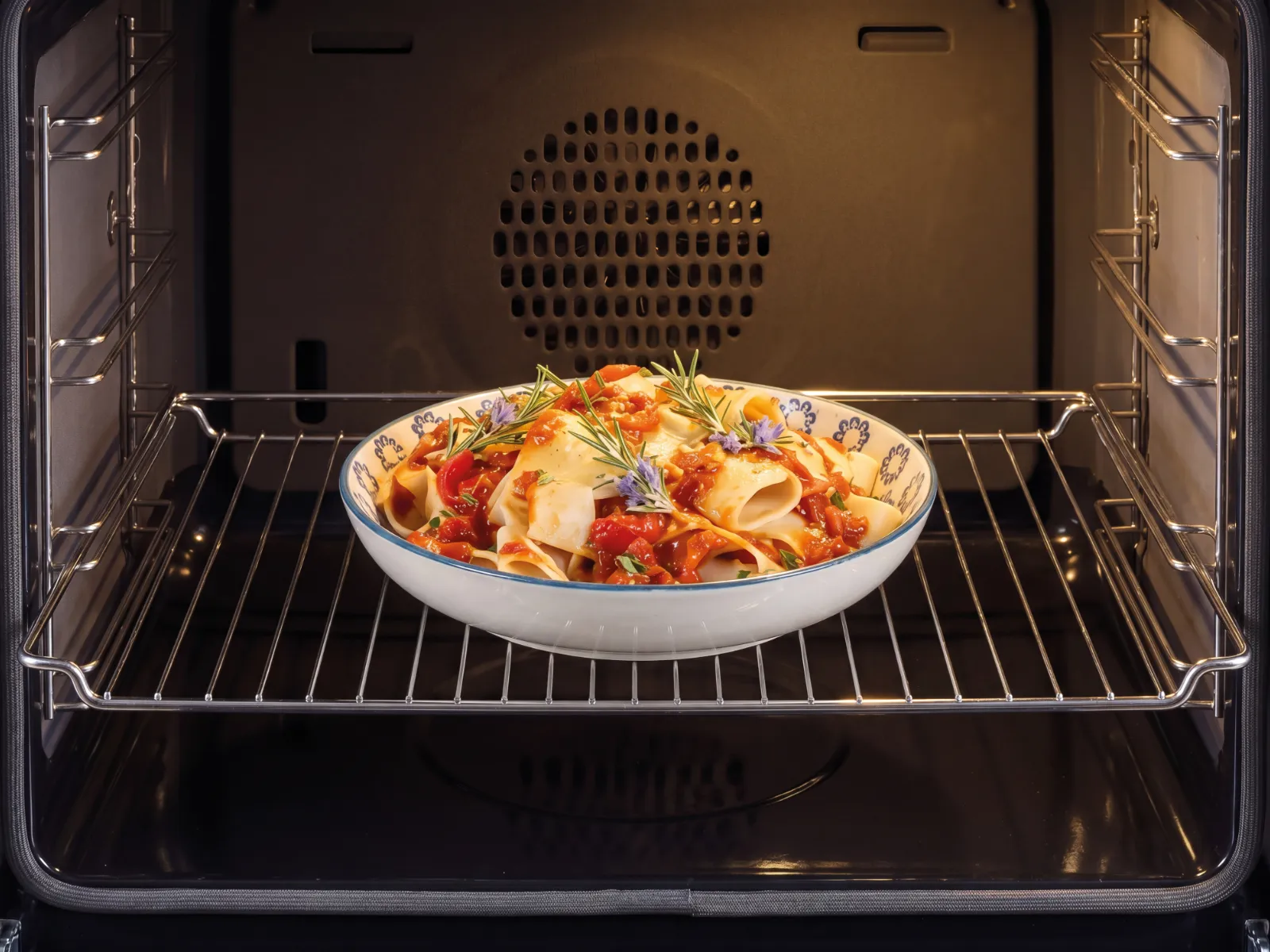 Eine Schüssel mit Pasta in Tomatensauce, garniert mit Rosmarin und Blüten, steht auf einem Gitterrost in einem leeren, hell beleuchteten modernen Backofen.