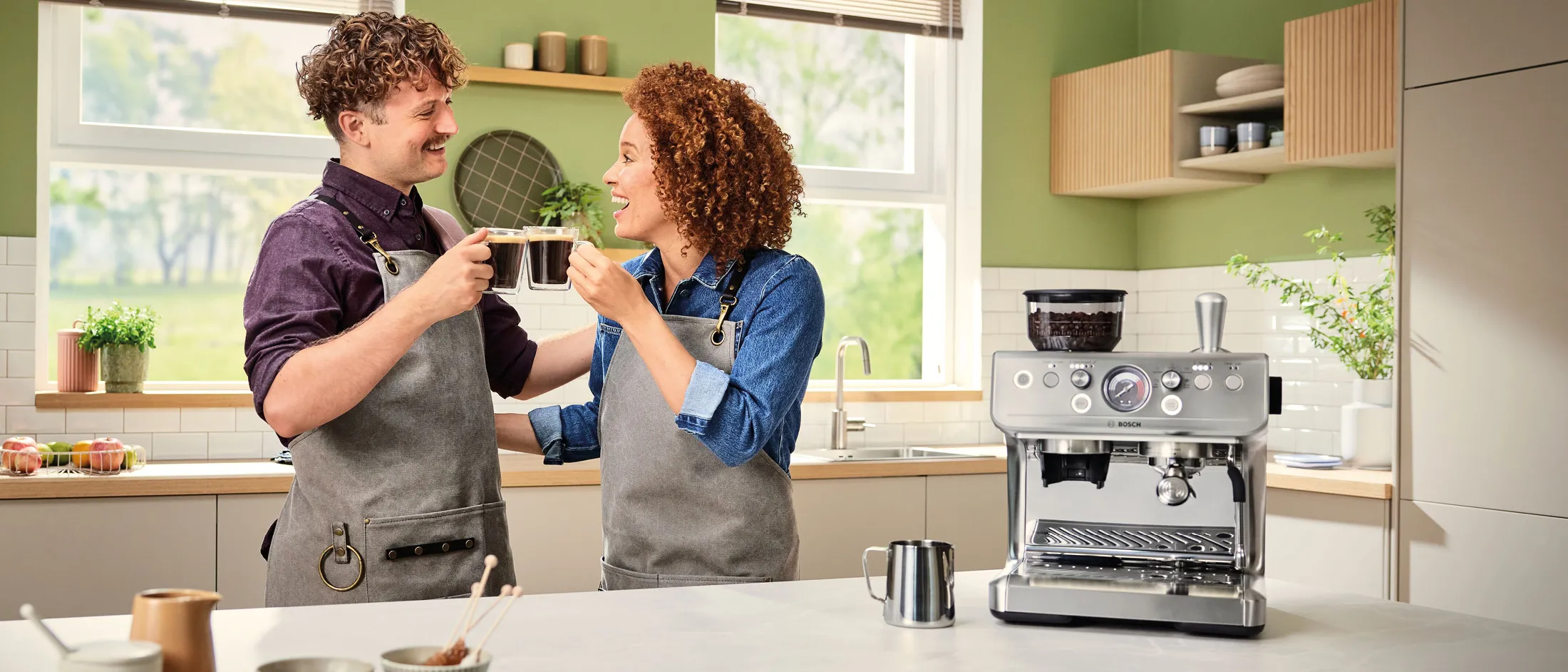 Deux baristas sourient et trinquent avec leurs tasses de café à côté d'une machine à expresso dans une cuisine moderne et lumineuse.