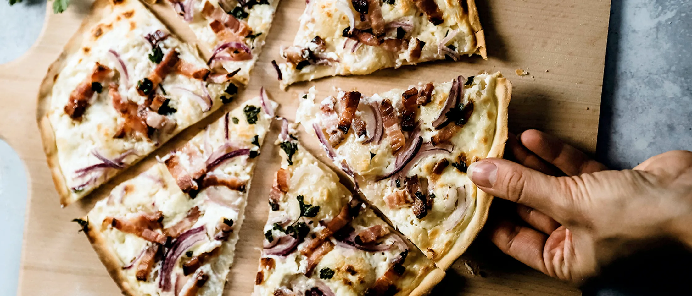 Hand greift nach einem Stück Flammkuchen mit Zwiebeln und Speck auf einem Holzbrett