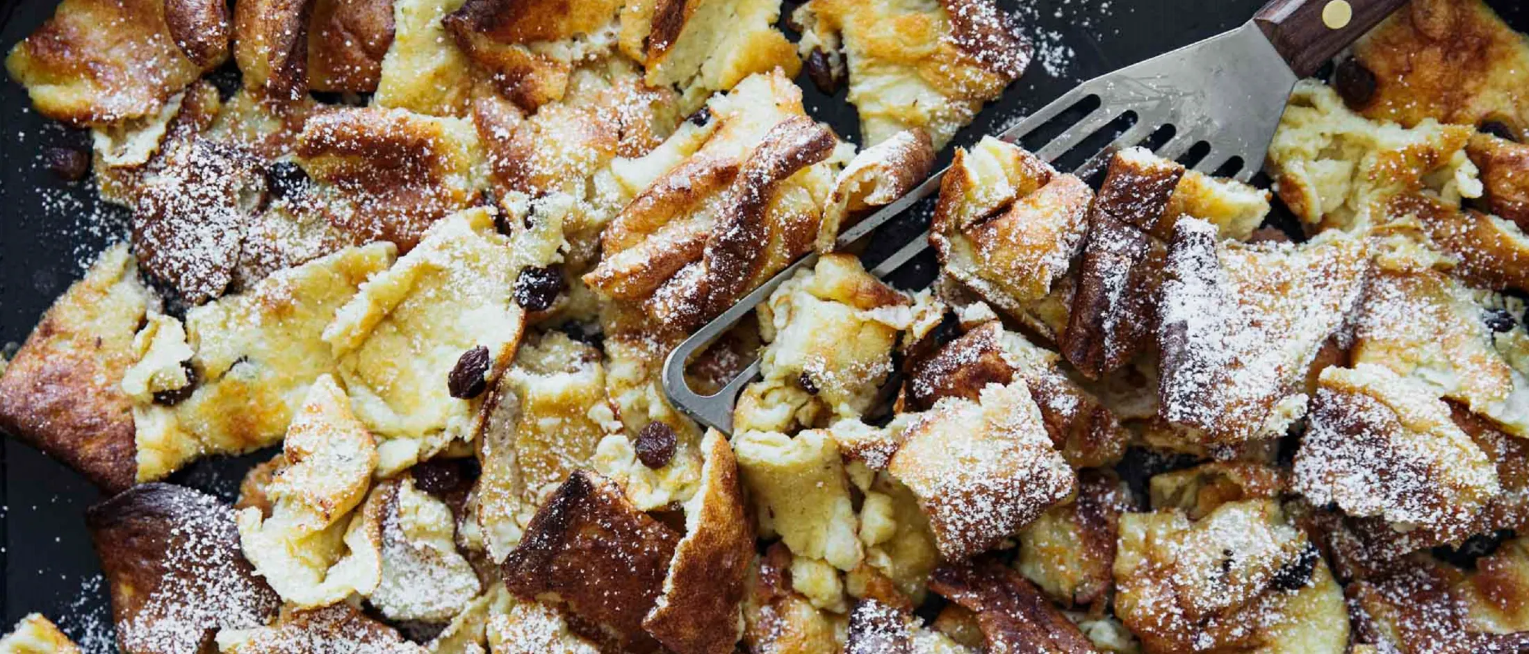 Nahaufnahme von zerzupftem Kaiserschmarrn mit Puderzucker auf dunklem Untergrund und einer Gabel