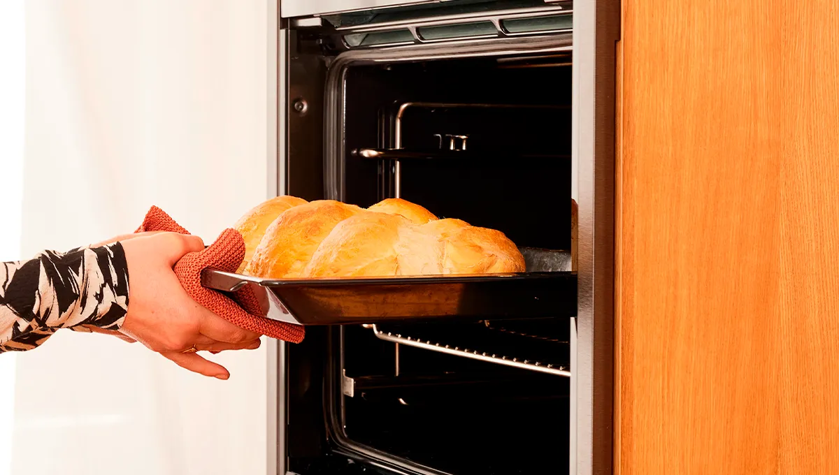 A close-up of hands in an oven mitt removing a tray of freshly baked, golden-brown bread from a stainless steel NEFF built-in oven.