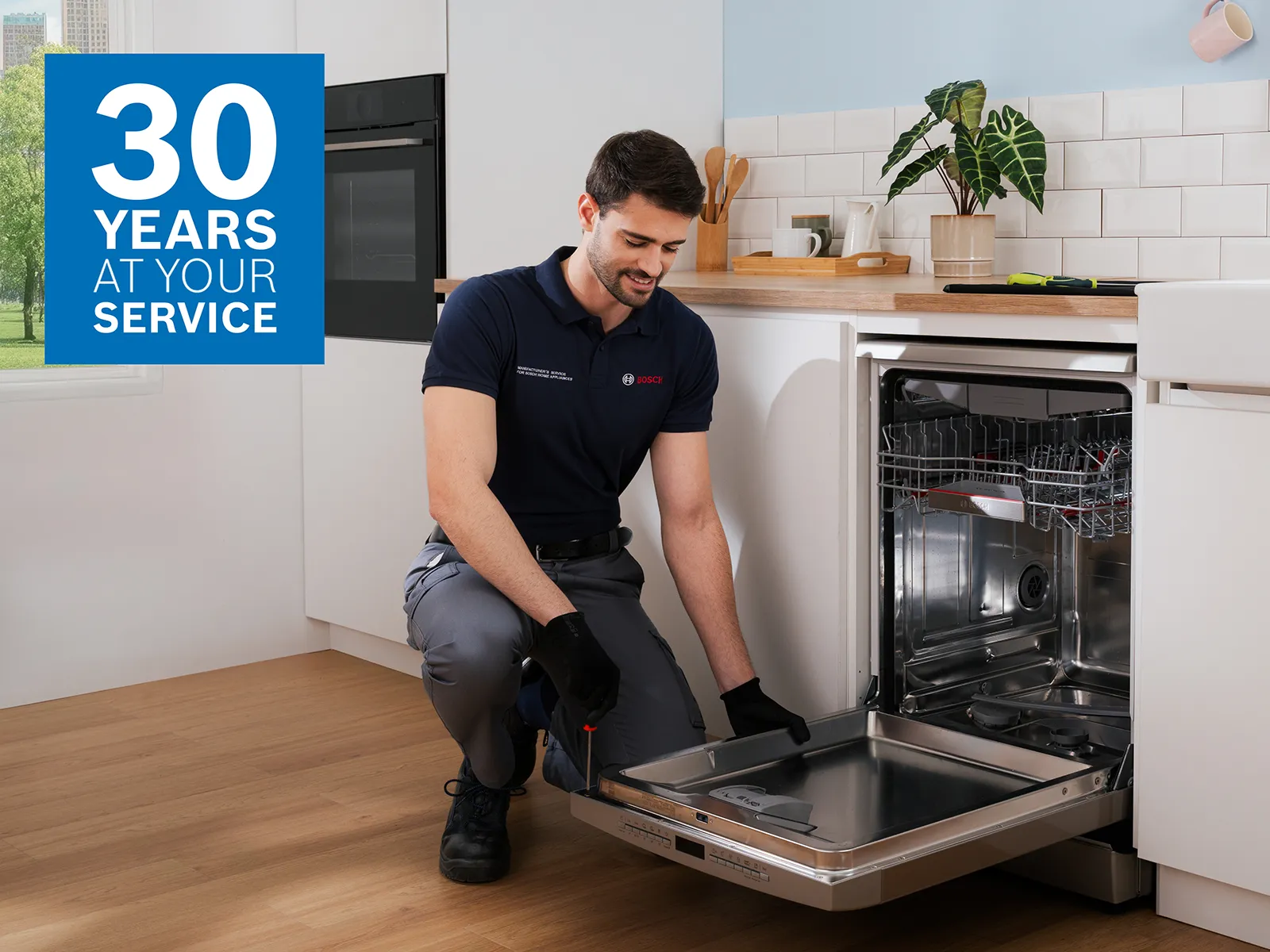 En Bosch-servicetekniker er ved at reparere en opvaskemaskine i et moderne køkken. I billedets venstre hjørne er teksten "30 YEARS AT YOUR SERVICE".