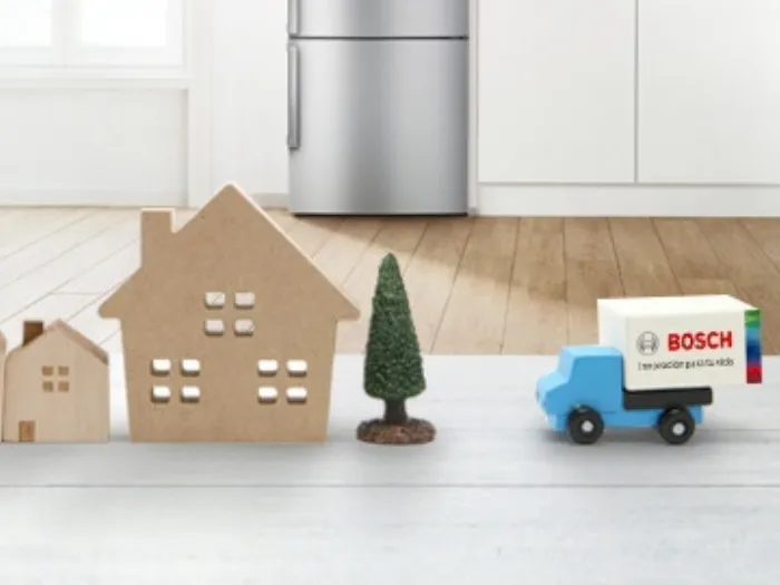 Casitas de cartón en miniatura y árboles de juguete verdes bordean una superficie blanca mientras un camión de reparto azul de Bosch atraviesa una cocina moderna y luminosa.
