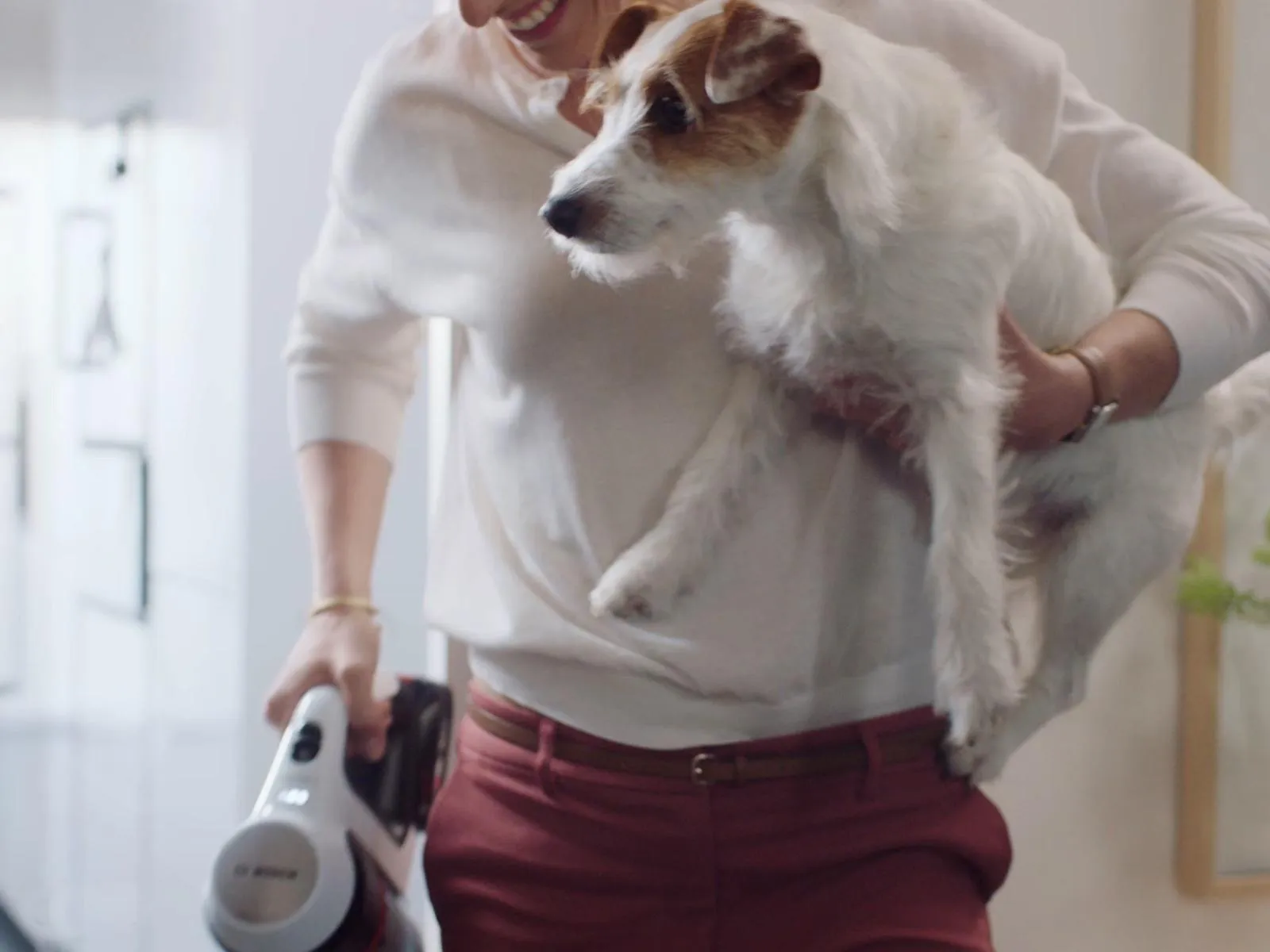 Person holding a small white and brown dog in one arm while carrying a handheld vacuum cleaner in the other hand.