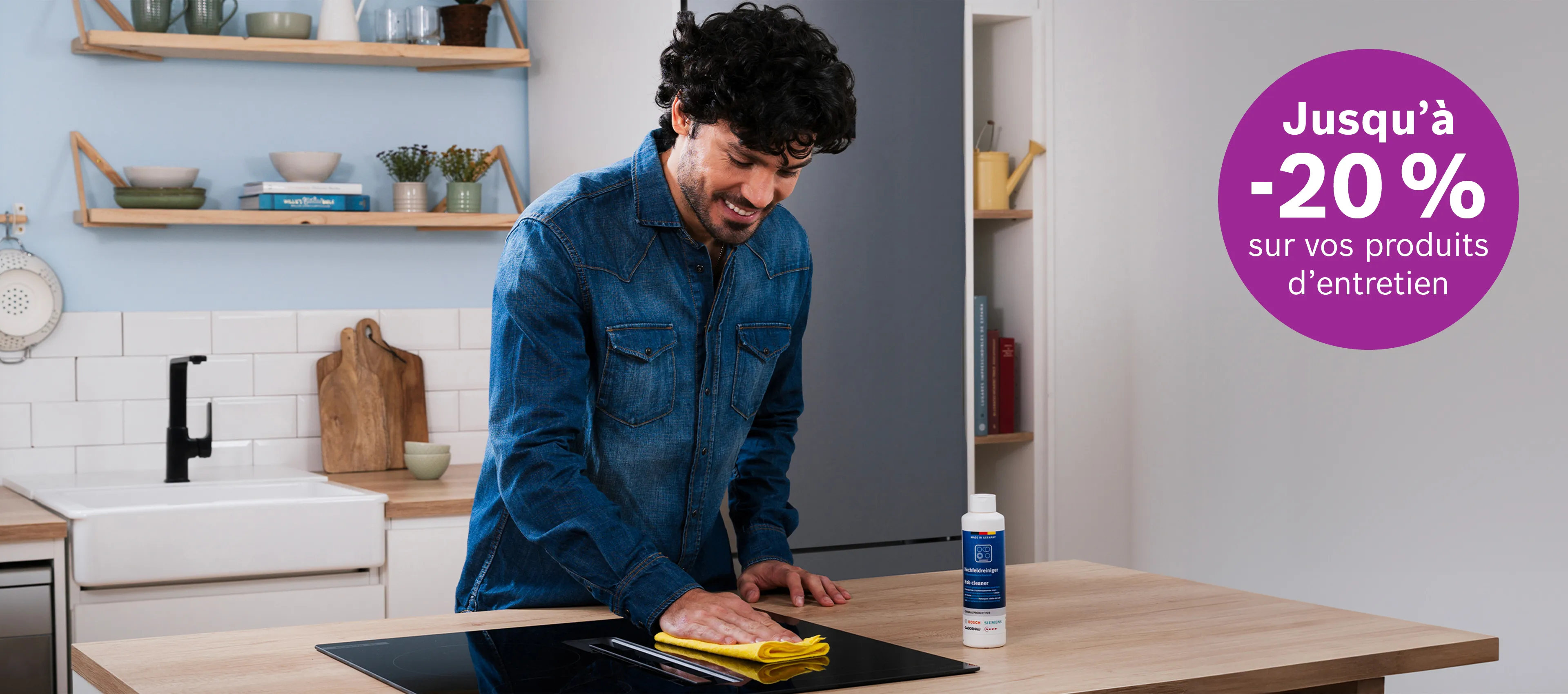 Homme souriant nettoyant une plaque de cuisson dans une cuisine moderne avec un autocollant indiquant 'Jusqu'à -20 % sur vos produits d'entretien'
