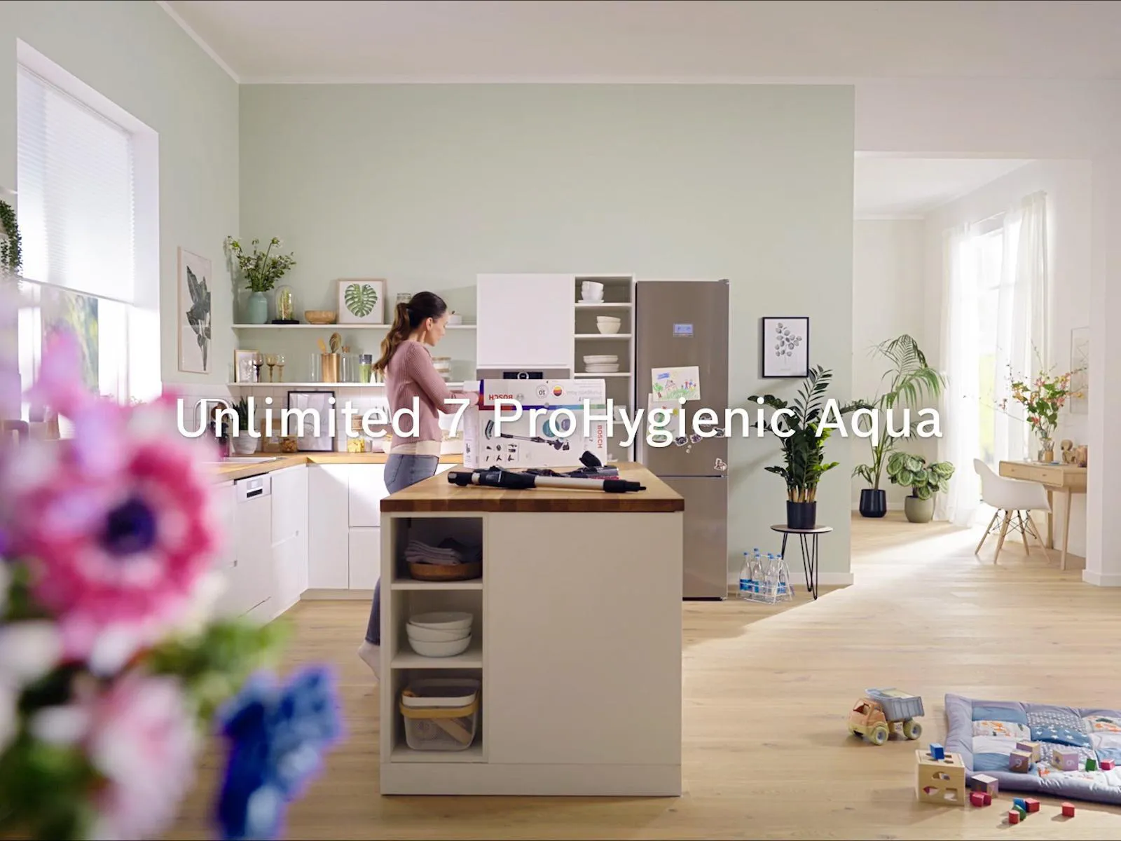 Modern kitchen with a woman preparing food, light wood flooring, light green walls, and toys scattered on the floor.
