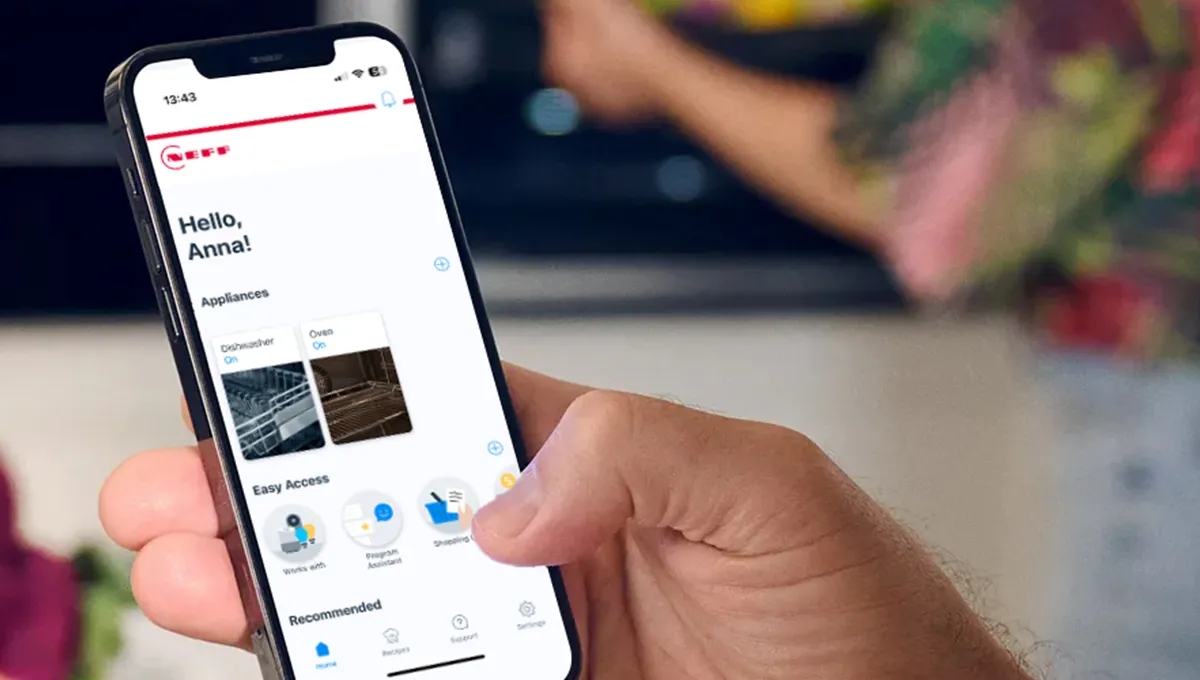 Κοντινό πλάνο σε ένα χέρι που κρατάει ένα smartphone το οποίο δείχνει την εφαρμογή Home Connect, ενώ κάποιος άλλος εργάζεται στην κουζίνα στο φόντο.