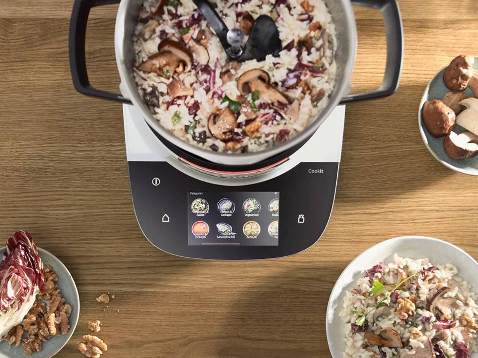 Der Bosch Cookit von oben mit einem Risotto im Topf. Das Display zeigt Automatikprogramme. Daneben Zutaten und fertige Teller.