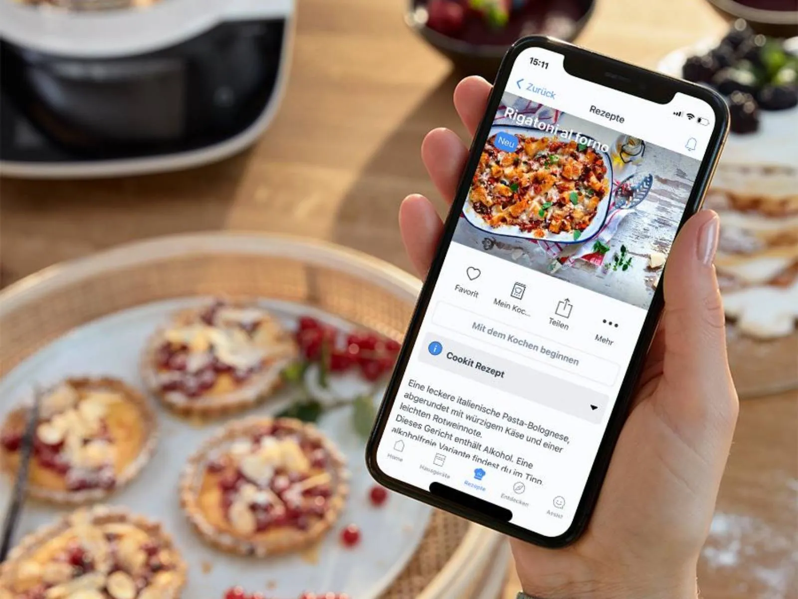 Eine Hand hält ein Smartphone mit einem Cookit Rezept in der Home Connect App. Im Hintergrund sind frisch gebackene Törtchen.