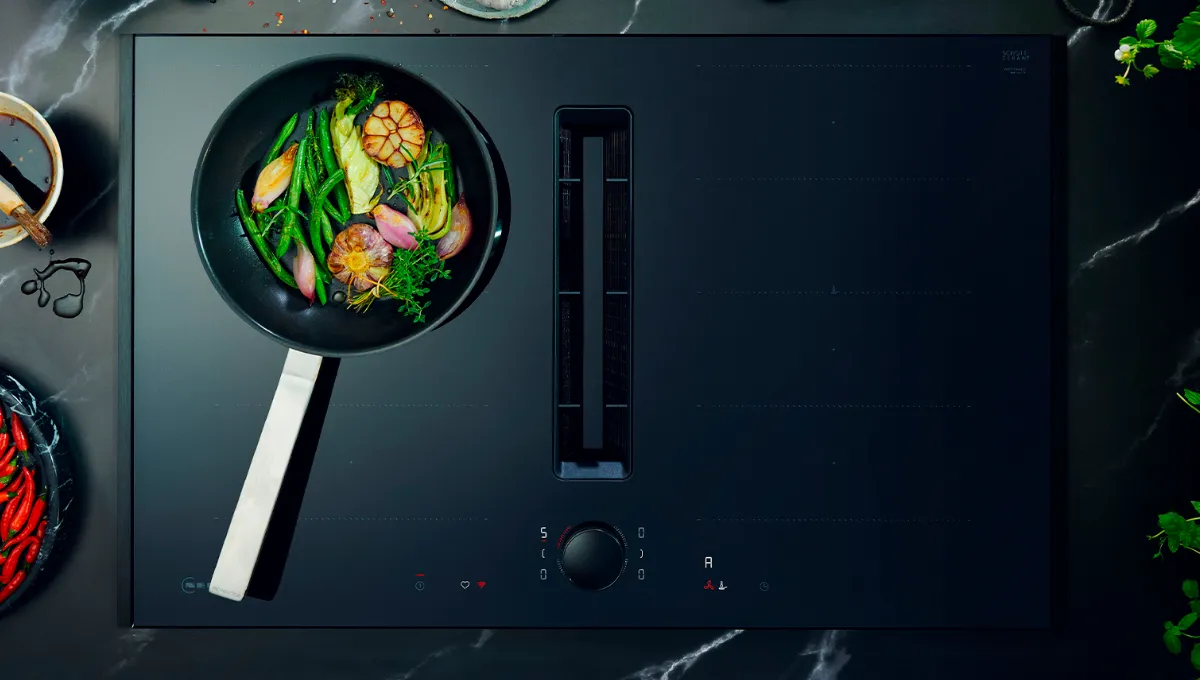Moderno piano cottura nero con una padella che cuoce fagiolini, aglio e scalogno, circondato da peperoncini freschi, bulbi d'aglio e spezie.