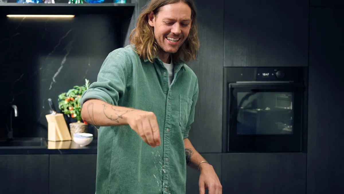 Uomo con camicia verde che sparge condimento su una teglia in una cucina moderna.