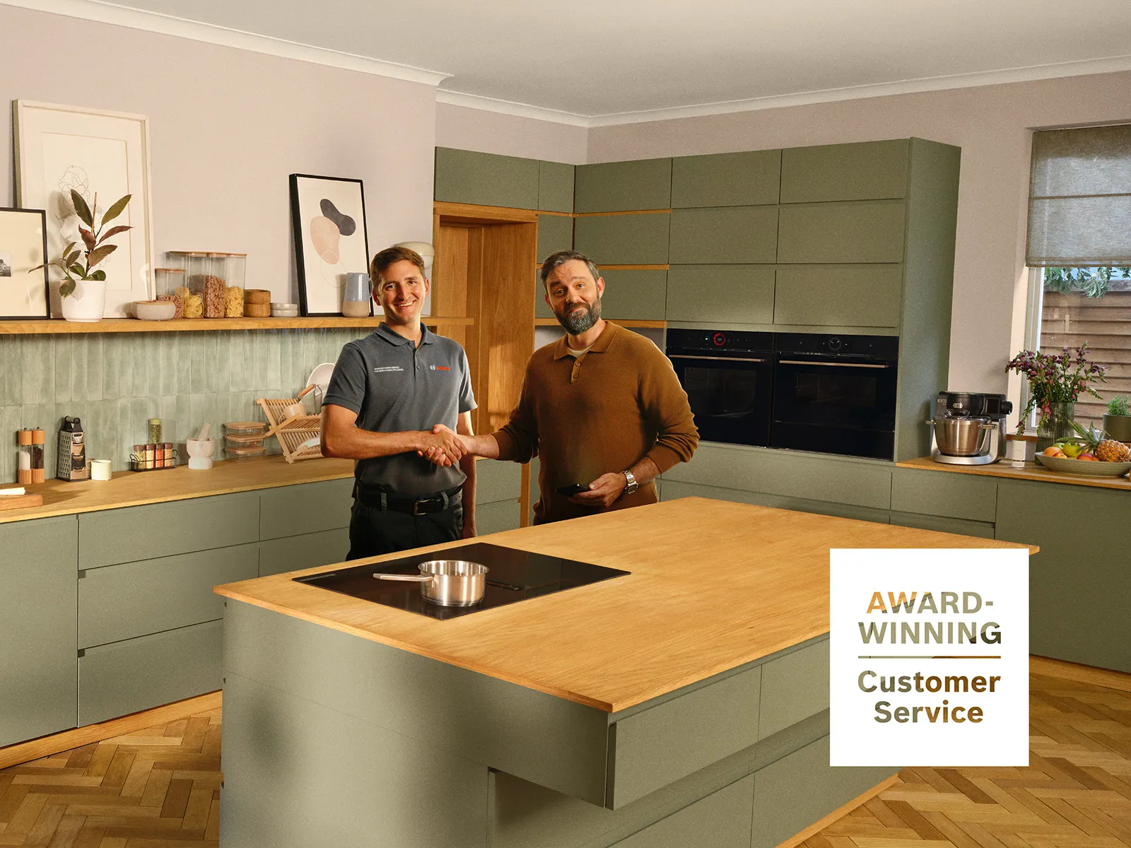 Two men standing in a modern olive-green kitchen with wooden countertops, built-in appliances, and Bosch kitchen equipment. A badge reads “Award Winning Customer Service.”