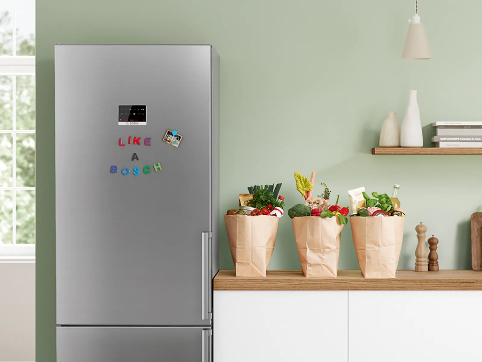 Frigorifero combinato Bosch in acciaio inox con la scritta magnetica "Like a Bosch" e buste della spesa con cibo fresco sul piano cucina.
