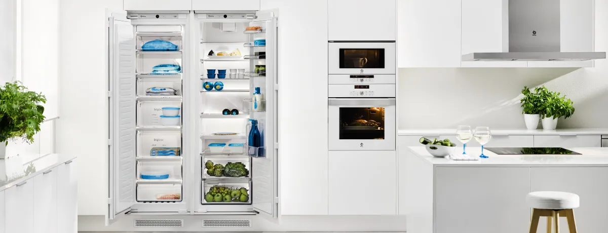 Cozinha moderna branca com frigorífico e congelador abertos e repletos de alimentos, forno encastrado, micro-ondas, plantas e ilha de cozinha com duas taças de vinho.