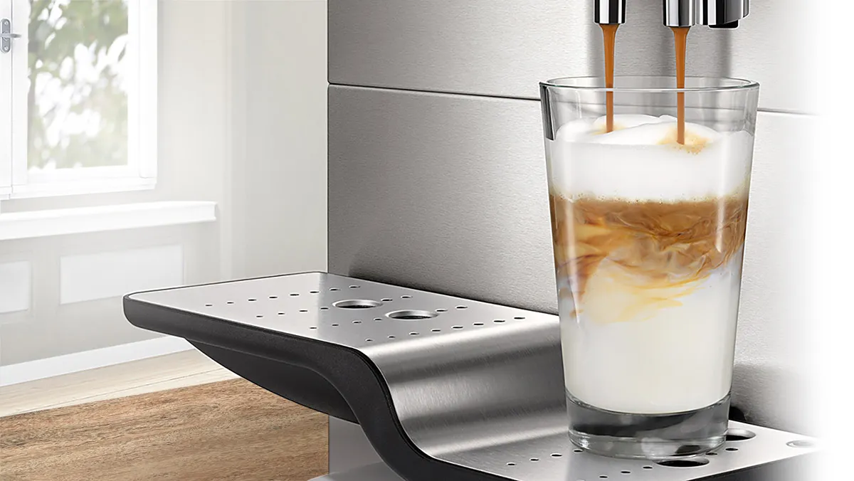 Eine Kaffeemaschine aus Edelstahl gießt Kaffee in ein Glas auf einer Holzoberfläche in der Nähe eines Fensters.