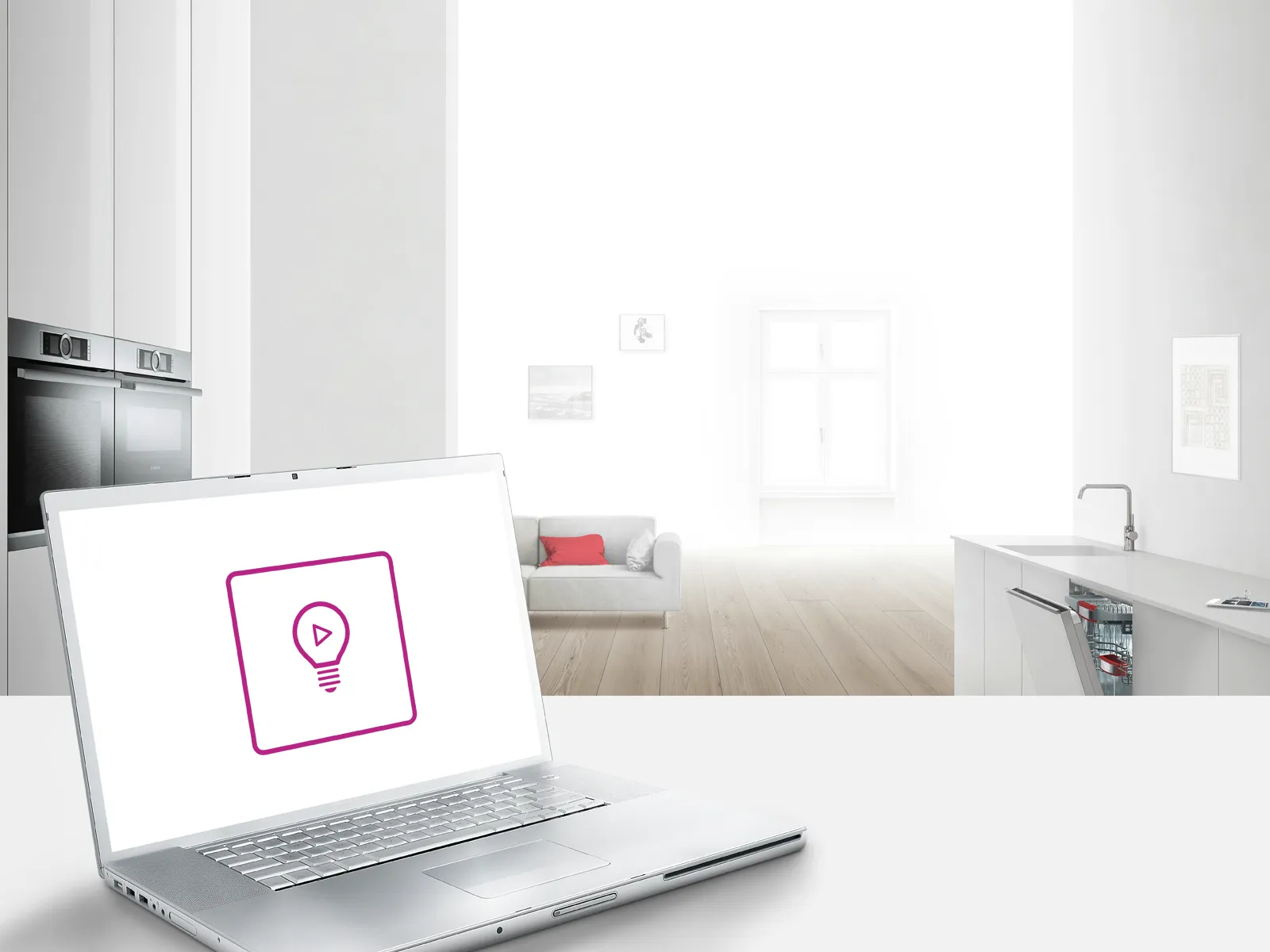 A laptop sits on a white surface in the foreground, displaying a lightbulb play icon, against a blurred background of a modern, white kitchen and living space.