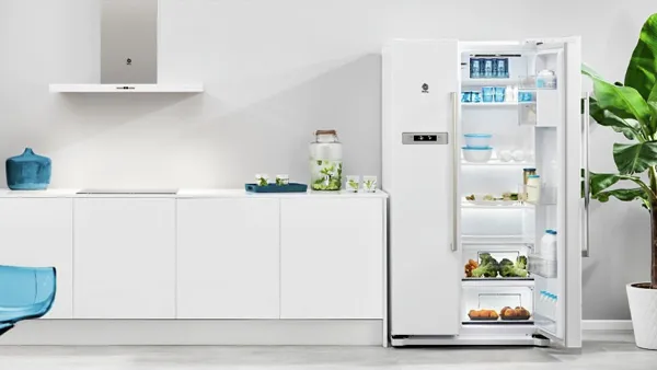 Cozinha moderna branca com um frigorífico aberto cheio de comida e bebidas, uma cadeira azul e uma planta verde em vaso.