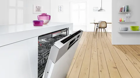Partially open integrated Bosch dishwasher built into a white kitchen island on a light wooden floor.